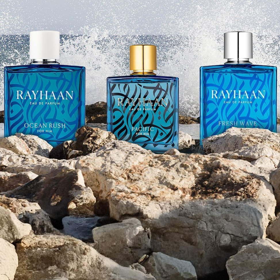 Three blue Rayhaan perfume bottles—Ocean Rush, Pacific, and Fresh Wave—stand on seaside rocks with ocean waves splashing behind them.