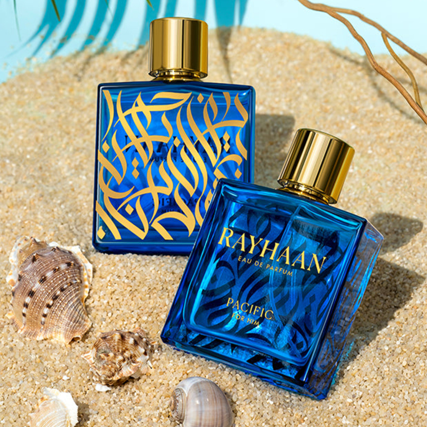 Two blue perfume bottles(font side and back side) with gold caps sit on sand beside seashells in a bright beach setting.