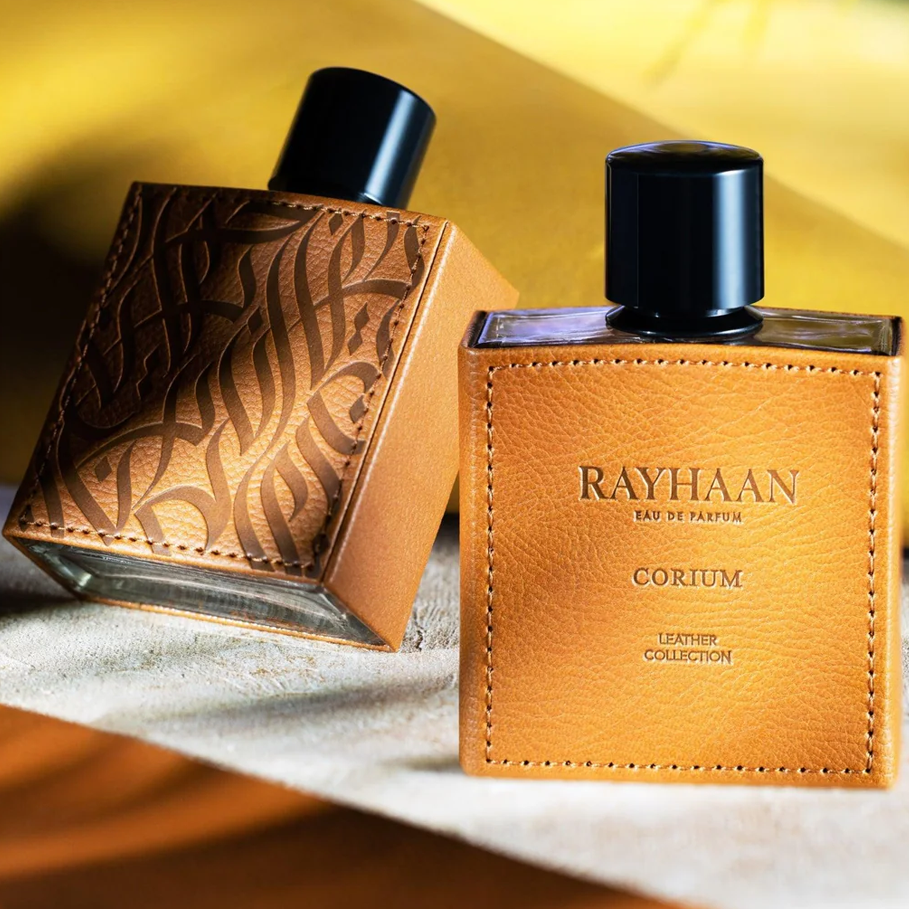 Two brown Rayhaan Corium perfume bottles from the Leather Collection, wrapped in stitched leather, sit on a warm golden surface under soft light.