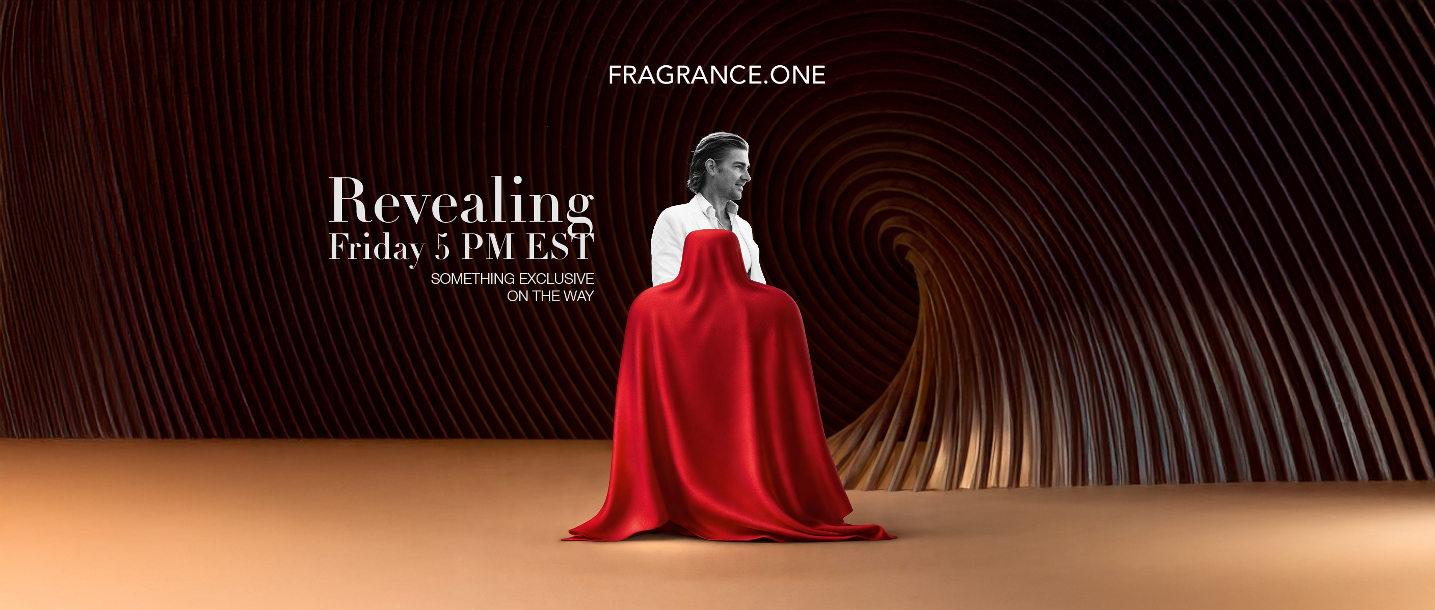 The image shows a modern, artistic tunnel made of curved wooden panels forming a dark, swirling backdrop. In the center stands a person dressed in a white shirt, shown in black and white, looking to the right with a confident expression. In front of them is a mysterious object completely covered by a flowing red cloth that drapes to the floor. Above, the text reads: “FRAGRANCE.ONE”. To the left of the person, large elegant text says: “Revealing Friday 5 PM EST”, followed by the smaller line “Something exclu