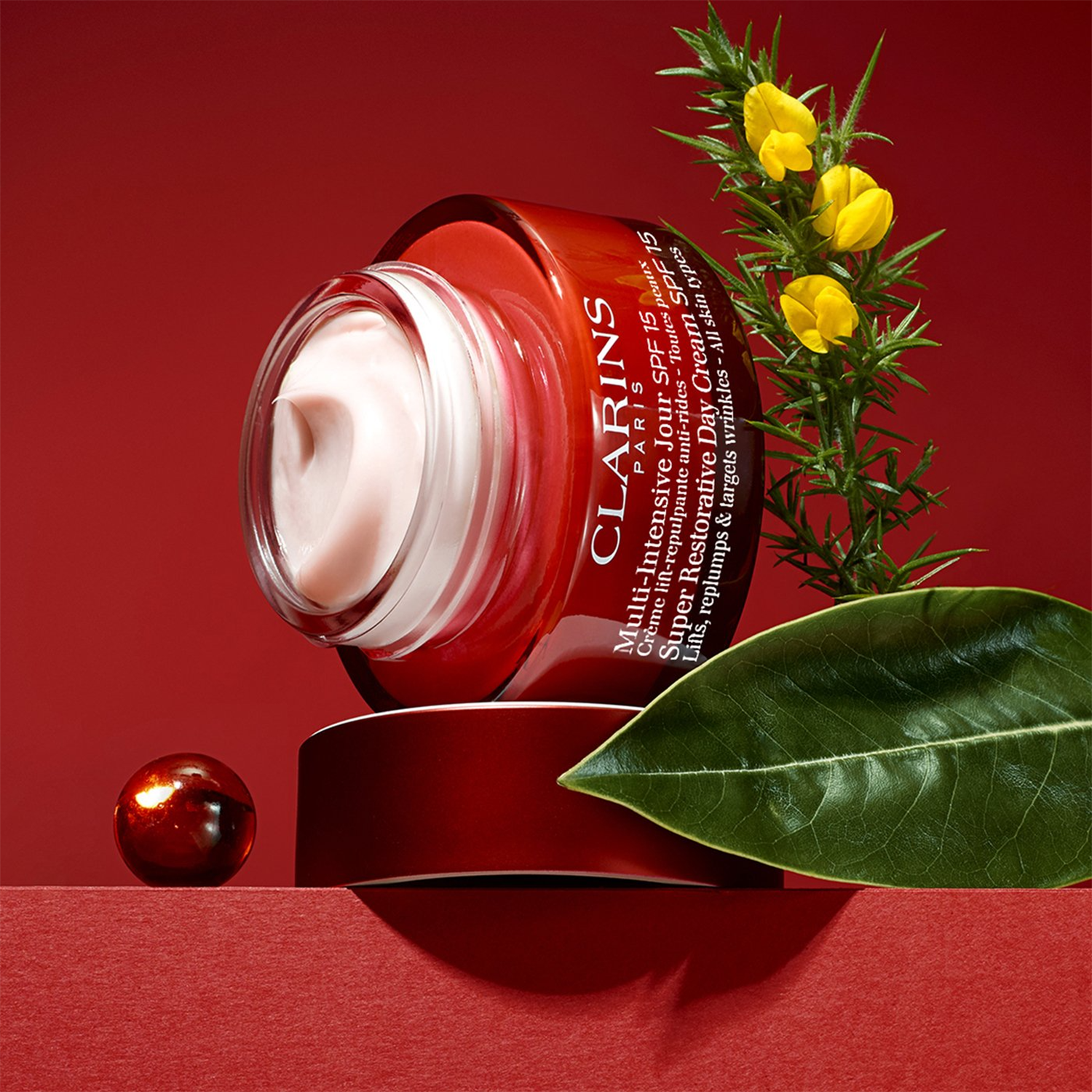Clarins Paris Super Restorative Day Cream in a red jar, open with cream visible, displayed against a red background with green leaves and yellow flowers.