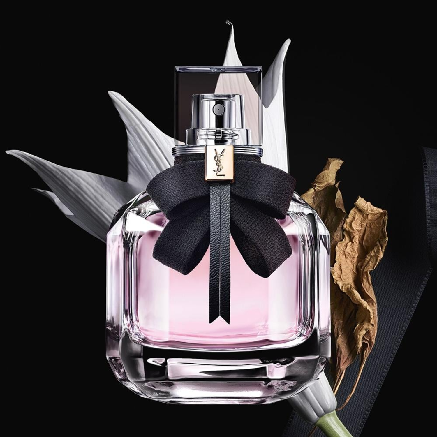 Yves Saint Laurent Mon Paris Eau de Parfum bottle with soft pink juice, crystal-clear glass, and a black ribbon bow, styled with a white flower and dried leaf on a black background.