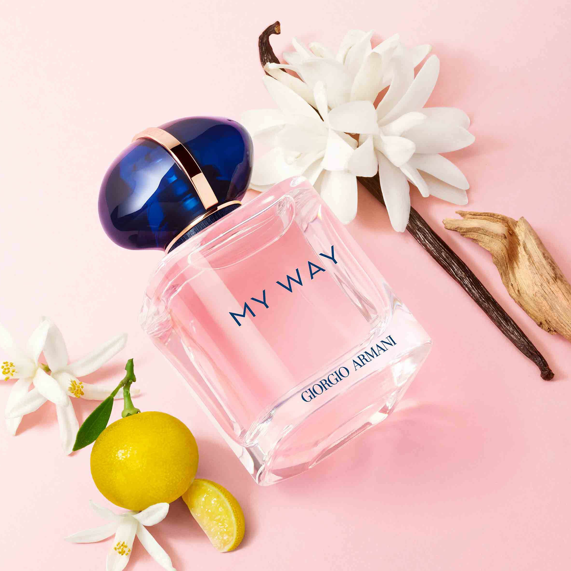 A Giorgio Armani My Way perfume bottle lies on a pink surface surrounded by white flowers, vanilla, wood, and lemon — symbolizing freshness and elegance.