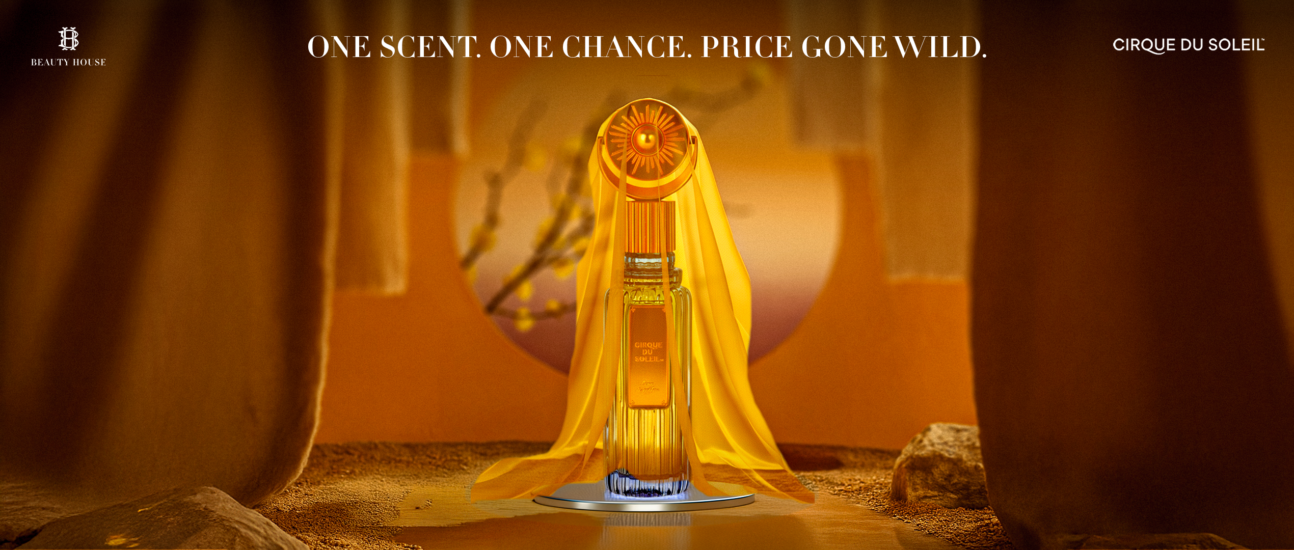 Here is a clear, short, accessibility-friendly visual description in paragraph form:

The image shows a dramatic golden scene with soft, flowing curtains on both sides, framing a perfume bottle in the center. The bottle is tall and elegant, glowing in shades of amber and gold. A sheer yellow fabric drapes over it like a cape, and the cap features a round sunburst design. The bottle sits on a small reflective platform on sandy ground, creating a warm, theatrical atmosphere. The text above reads, “One scent. 